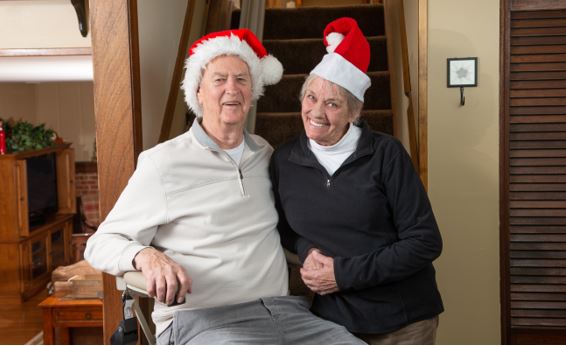 senior man and woman wearing Santa hats in home while man rides stairlift